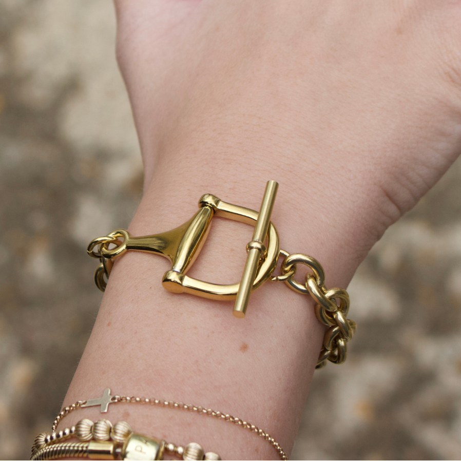 A close-up of a person's wrist wearing a gold horse-bit style chain bracelet with a toggle clasp, layered with delicate gold chains and a ring with a heart cutout.