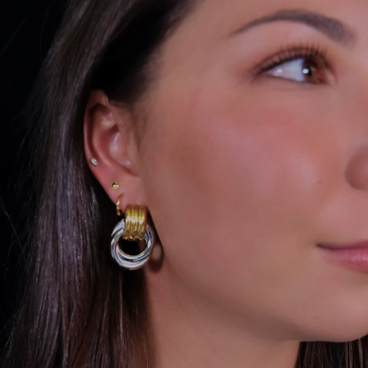 Close-up of a woman wearing gold hoop earrings and a gold necklace with a horse bit pendant against a black background.