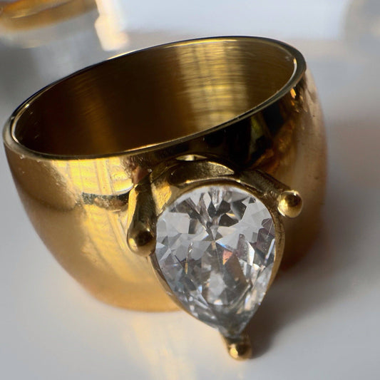 A wide gold ring featuring a large, teardrop-shaped clear gemstone in a prong setting, resting on a reflective white surface.