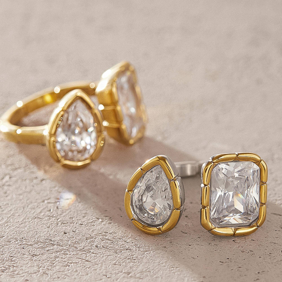 Gold jewelry set featuring a ring and matching stud earrings with clear teardrop and rectangular gemstones, displayed on a textured beige surface.