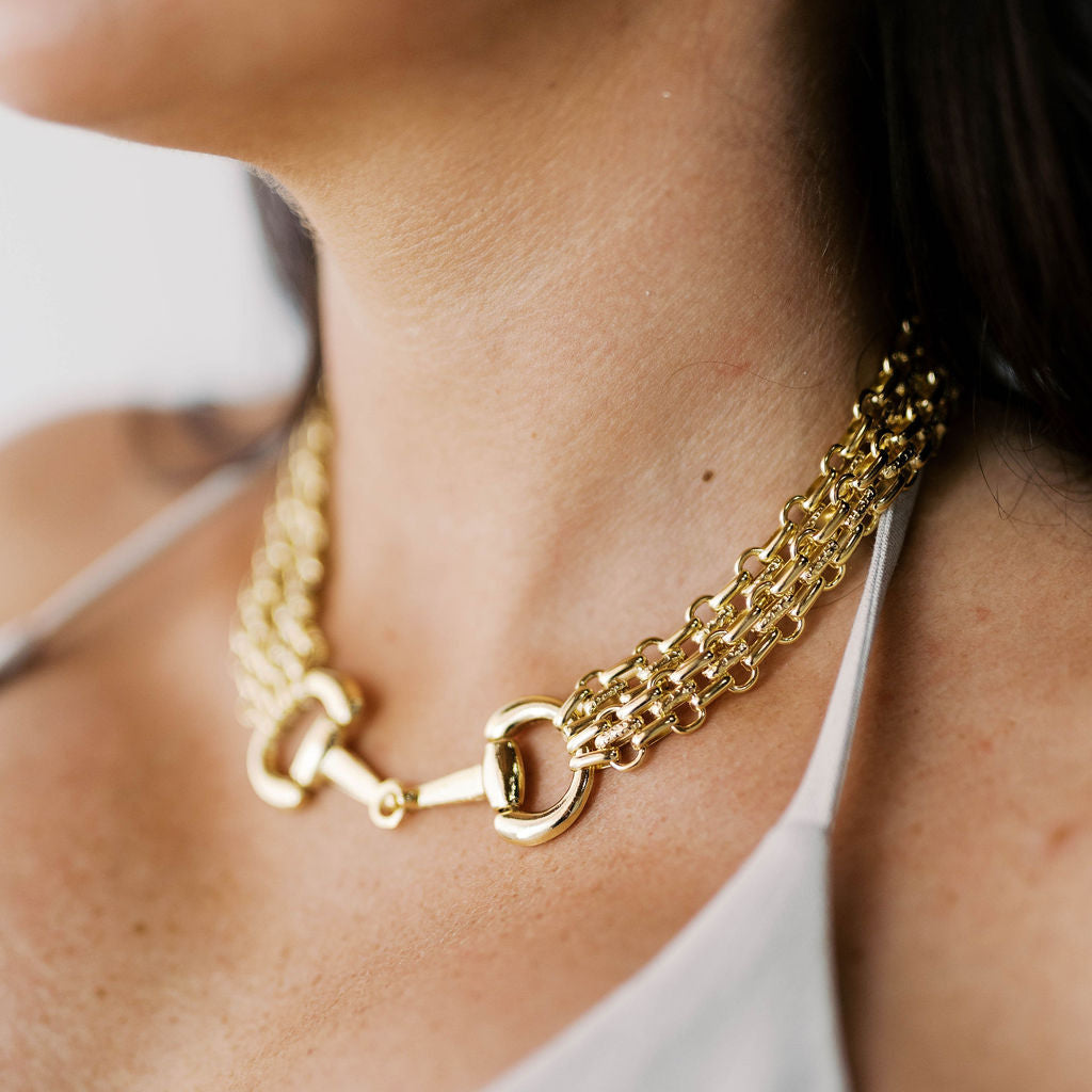 A close-up lifestyle shot of a model wearing a gold horsebit chain necklace over a white tank top.